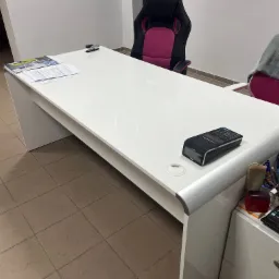 Białe biurko z dokumentami, zszywaczem, kalkulatorem i terminalem płatniczym w biurze, widoczny fotel gamingowy i szafka z przyborami biurowymi.