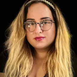 Portret kobiety z blond włosami, w okularach i z ozdobną opaską na głowie, na czarnym tle.