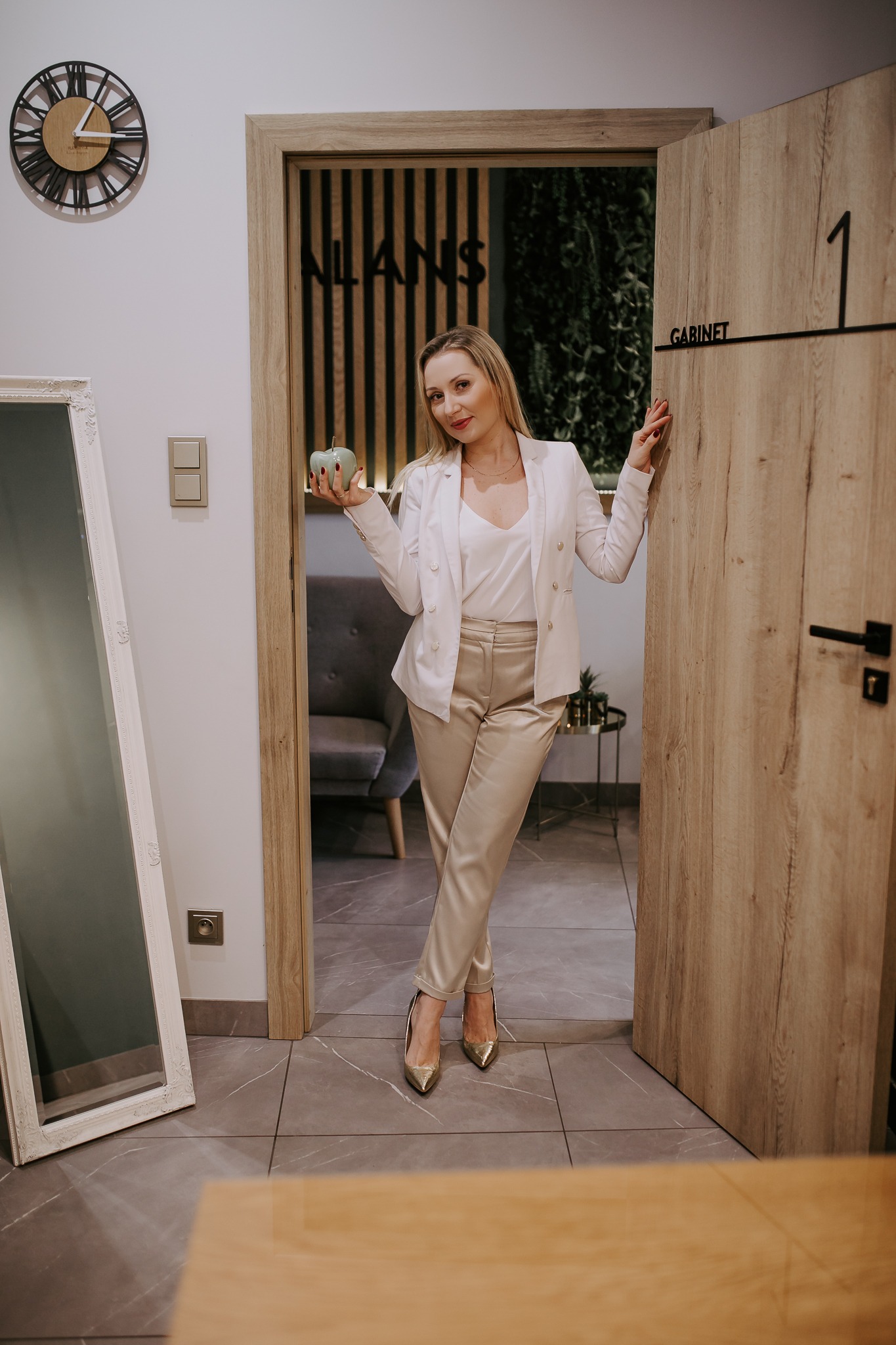 Elegancka blondynka w białym garniturze pozuje w drzwiach gabinetu, trzymając w dłoni zielone jabłko, widoczny zegar na ścianie i lustro w ozdobnej ramie.
