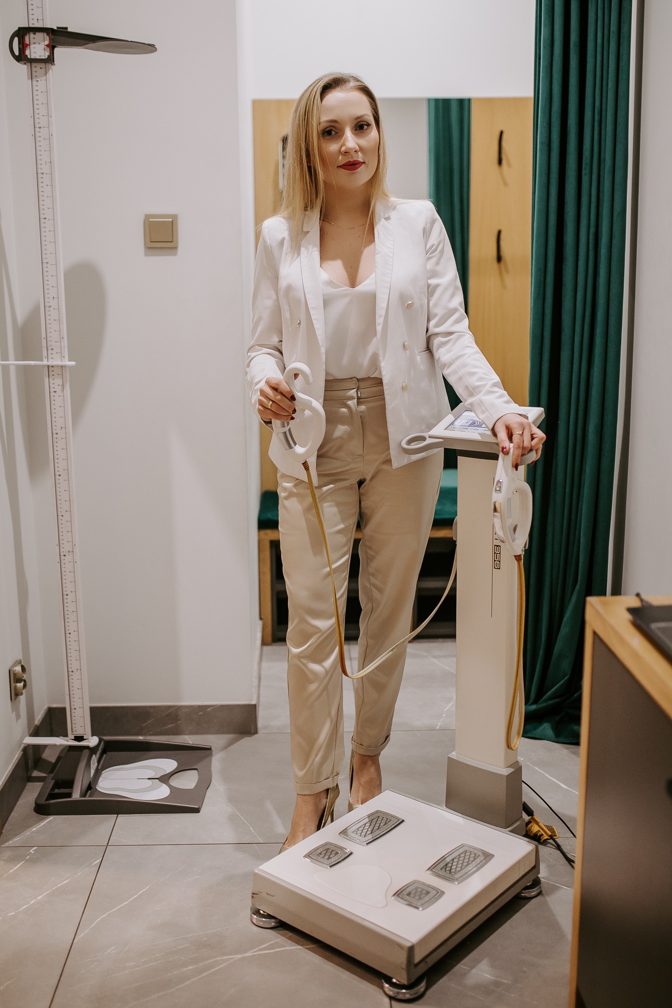 Dietetyk prezentuje analizator składu ciała z uchwytami, obok miarka wzrostu, kobieta w białym żakiecie.