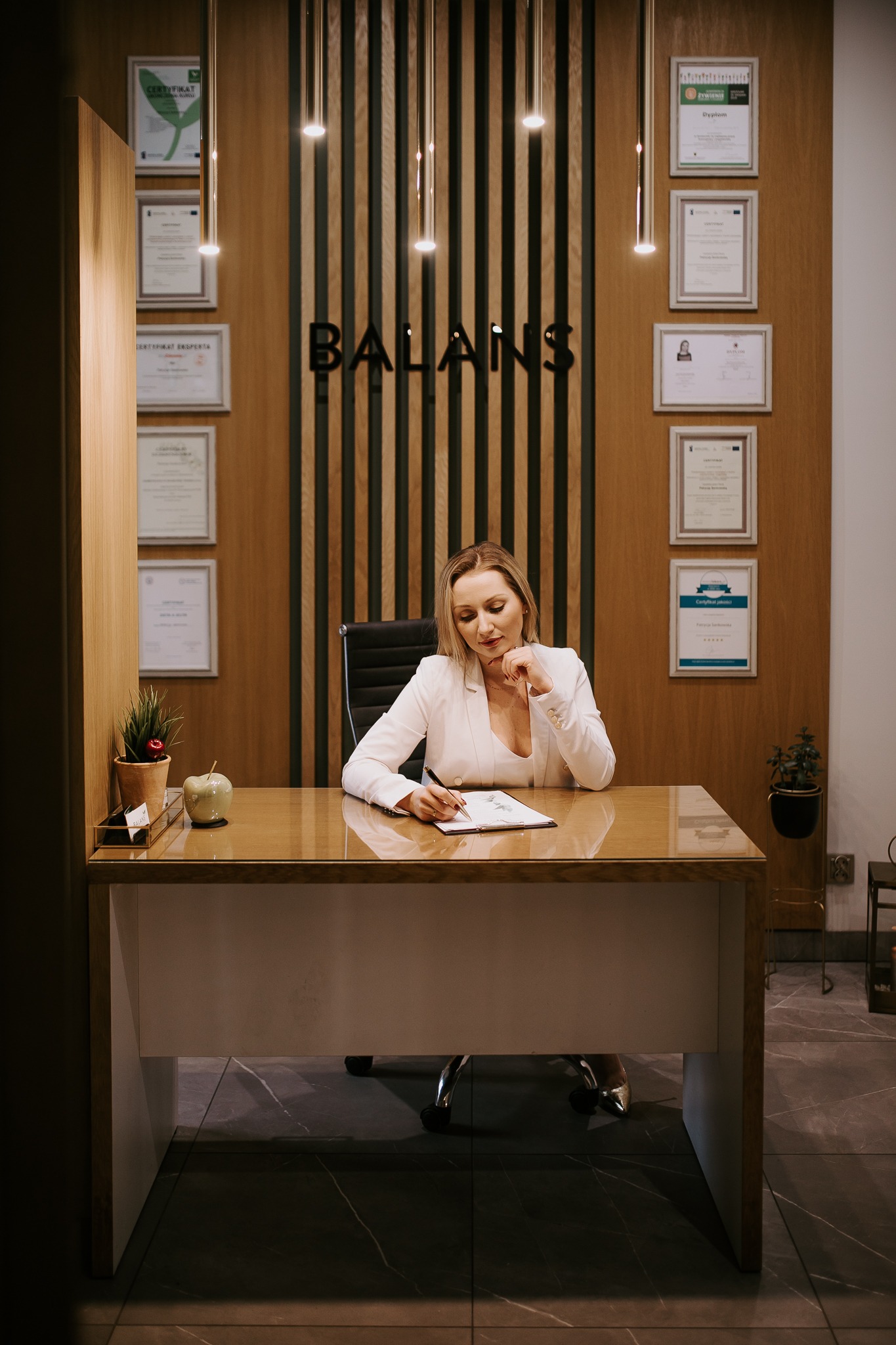 Kobieta w białym żakiecie siedzi za biurkiem w gabinecie, pisząc na kartce; w tle ściana z certyfikatami i napisem 'BALANS'.