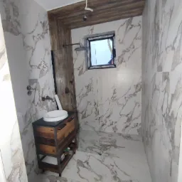 Nowoczesna łazienka z marmurowymi płytkami, drewnianą obudową prysznica i szafką pod umywalkę w stylu industrialnym, widoczne niedokończone podłączenie oświetlenia.