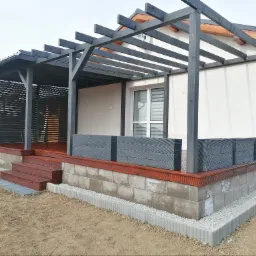 Zadaszony taras z drewnianą pergolą pomalowaną na grafitowo, podest z drewna w kolorze mahoniu, obudowany szarymi betonowymi bloczkami i obrzeżem z kostki.