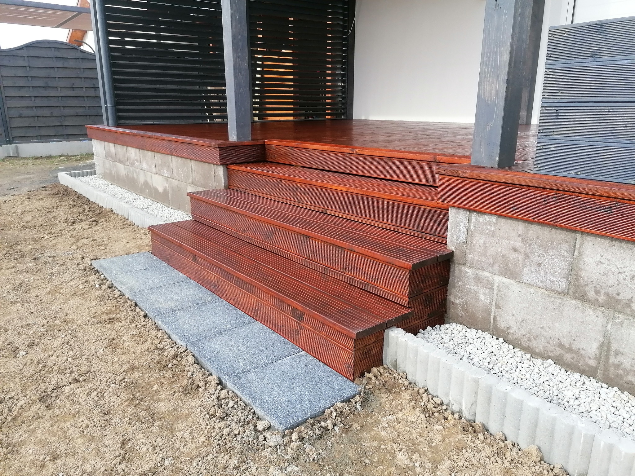 Nowoczesny taras z drewna kompozytowego w odcieniach brązu, z widocznymi schodami i obrzeżem z szarych płyt betonowych, otoczony jasnym żwirem i ziemią.