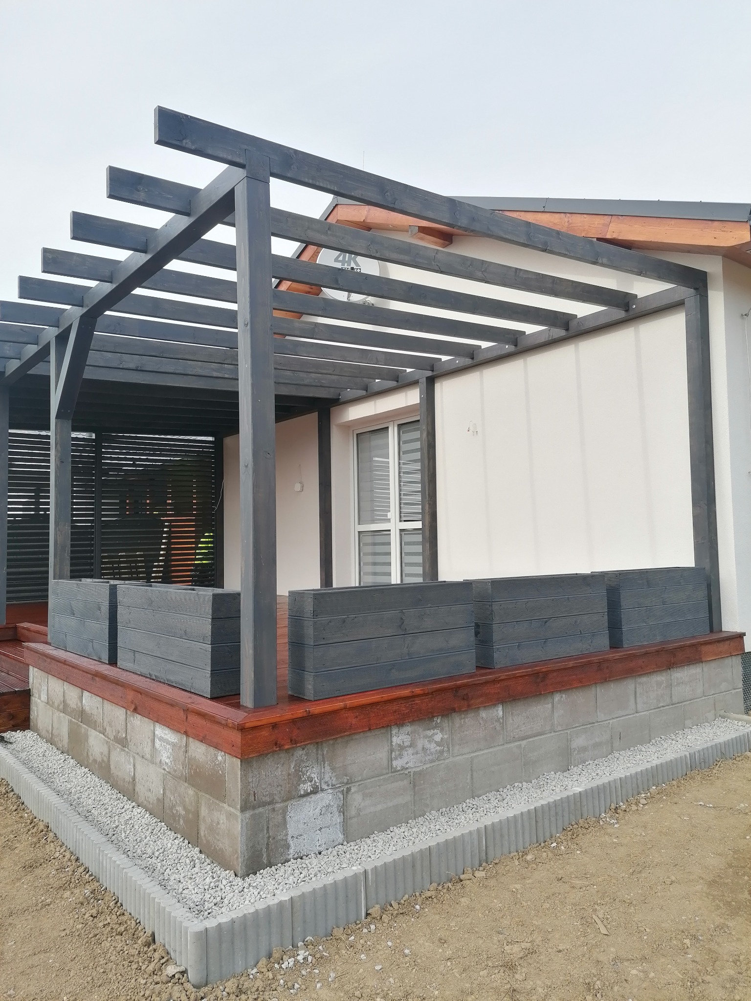 Zadaszony taras z drewnianą pergolą pomalowaną na szaro, obudowany donicami z tego samego drewna, z widoczną podmurówką z szarych bloczków betonowych i ozdobnym obrzeżem z kamyków.