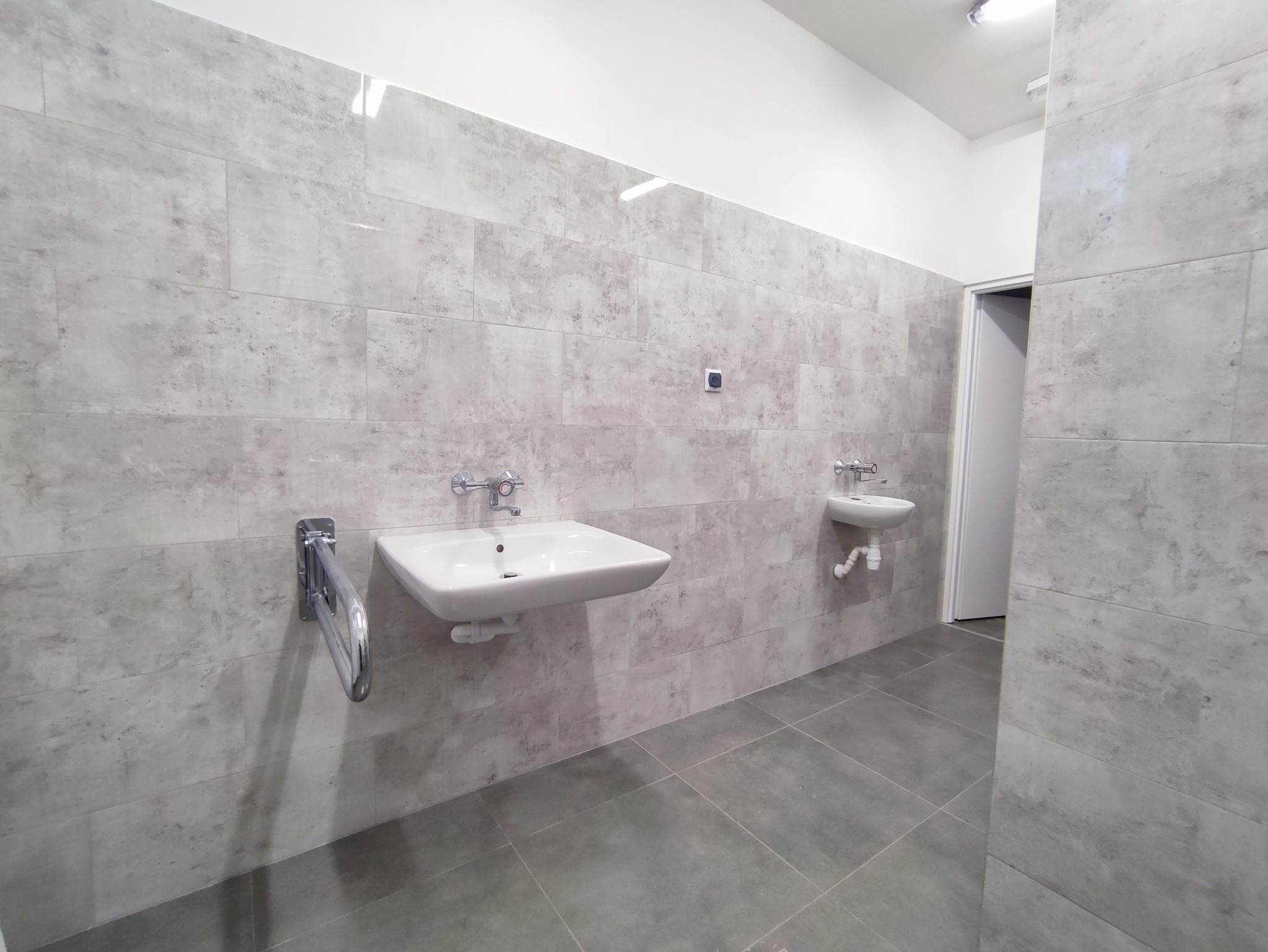 Wykończona łazienka z szarymi płytkami imitującymi kamień, białą umywalką z chromowaną baterią i uchwytem dla osób niepełnosprawnych. Na ścianie widoczny włącznik światła.