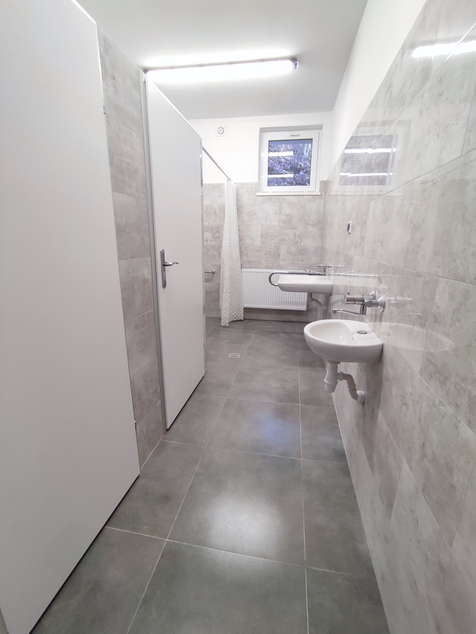 Publiczna toaleta z kabinami, umywalkami i szarą glazurą na ścianach i podłodze, widok z perspektywy wejścia.