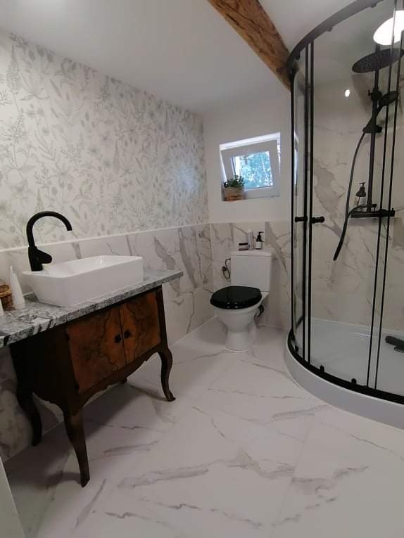 Umywalka nablatowa na stylizowanej szafce z ciemnego drewna, kabina prysznicowa z czarnymi profilami, toaleta z czarną deską, marmurowe płytki na podłodze i ścianach, oraz drewniana belka stropowa...