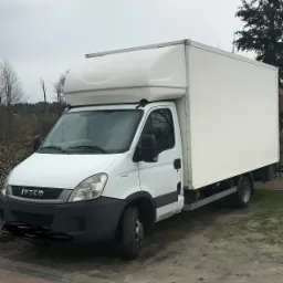 Biały samochód dostawczy Iveco z zabudową kontenerową, zaparkowany na nierównym terenie z widocznymi drzewami w tle, sfotografowany w pochmurny dzień.