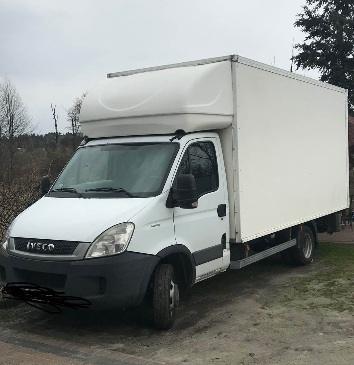 Biały samochód dostawczy Iveco z zabudową kontenerową, zaparkowany na nierównym terenie z widocznymi drzewami w tle, sfotografowany w pochmurny dzień.