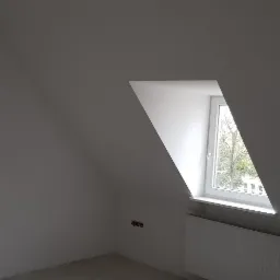 Poddasze ze skośnym oknem po malowaniu na biało, widoczny grzejnik i wystające kable ze ściany.