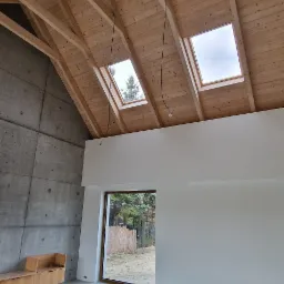 Wnętrze domu w stylu skandynawskim z drewnianym stropem z widocznymi belkami, betonową ścianą, dużym oknem z widokiem na zieleń i surową podłogą.