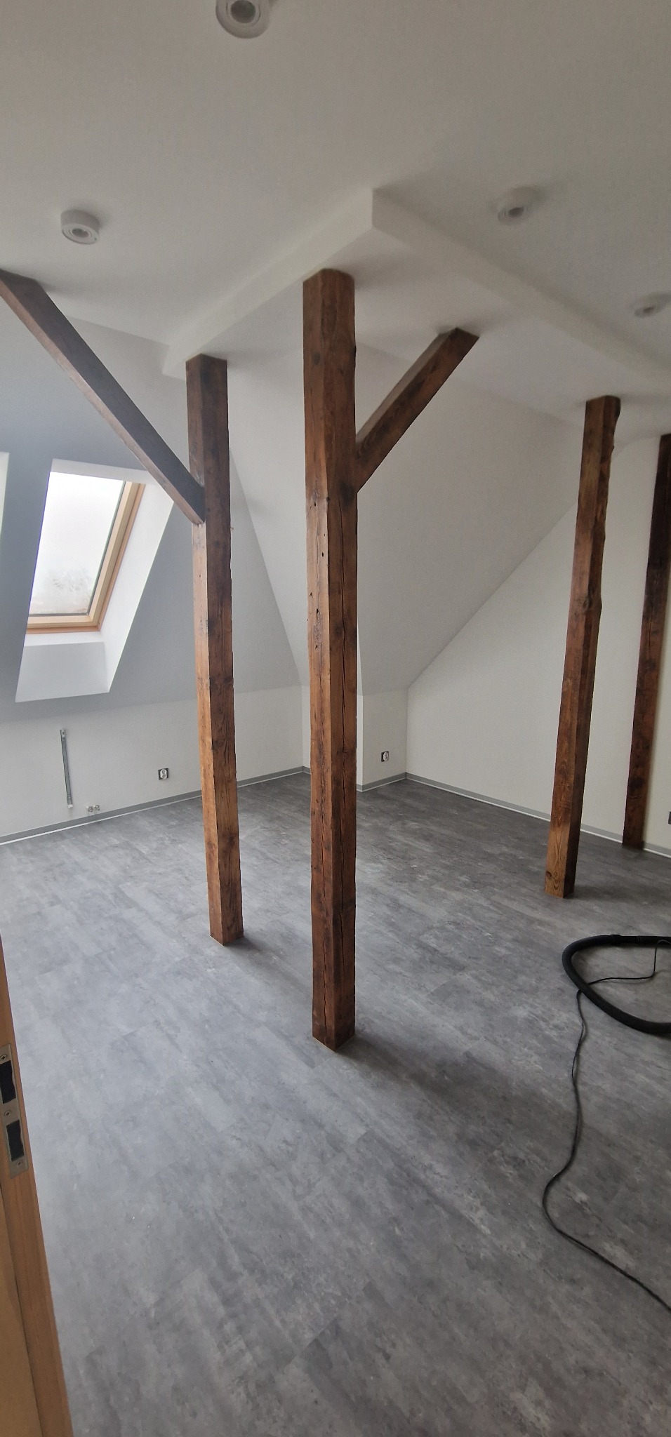 Poddasze po remoncie z drewnianymi belkami konstrukcyjnymi, szarą podłogą imitującą beton i widocznym oknem dachowym.