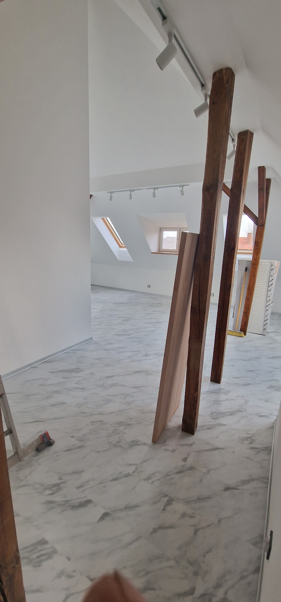 Poddasze w Gliwicach w trakcie wykończenia: widoczne drewniane słupy konstrukcyjne, marmurowe płytki podłogowe, białe ściany i sufit z oświetleniem punktowym. Częściowo widoczna drabina i narzędzia.