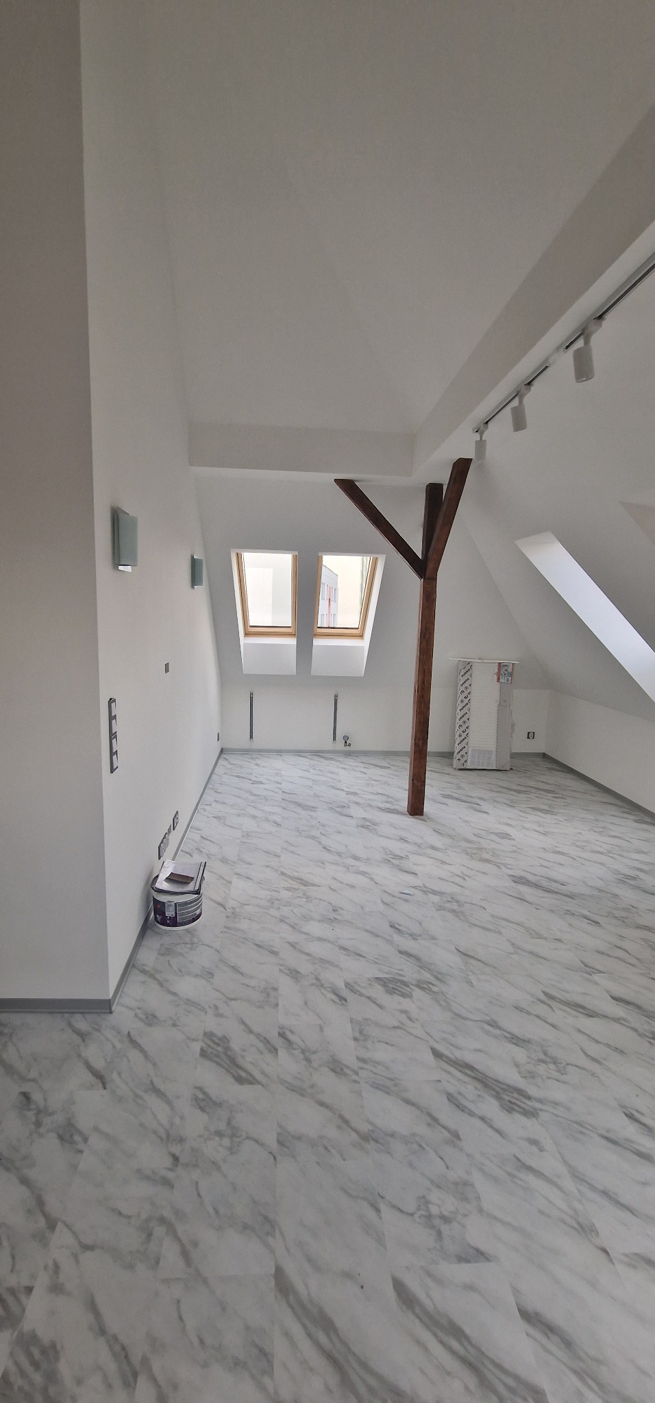 Poddasze po remoncie z marmurową podłogą, drewnianą konstrukcją nośną i nowymi oknami dachowymi. Wiadro z farbą przy ścianie wskazuje na świeże malowanie.