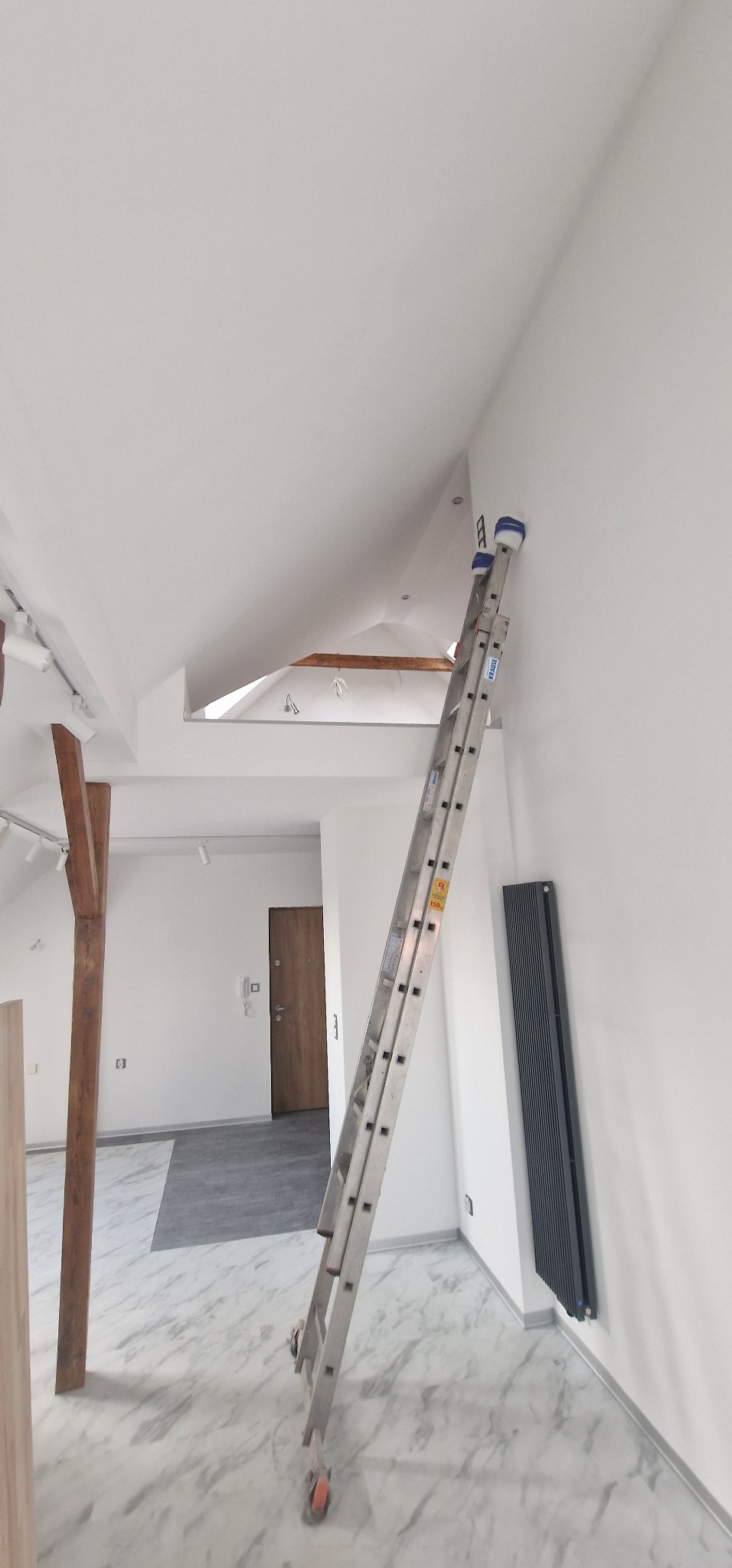 Wnętrze po remoncie, z białymi ścianami i marmurową podłogą, aluminiowa drabina oparta o ścianę, widoczny grzejnik panelowy i drewniany słup konstrukcyjny.