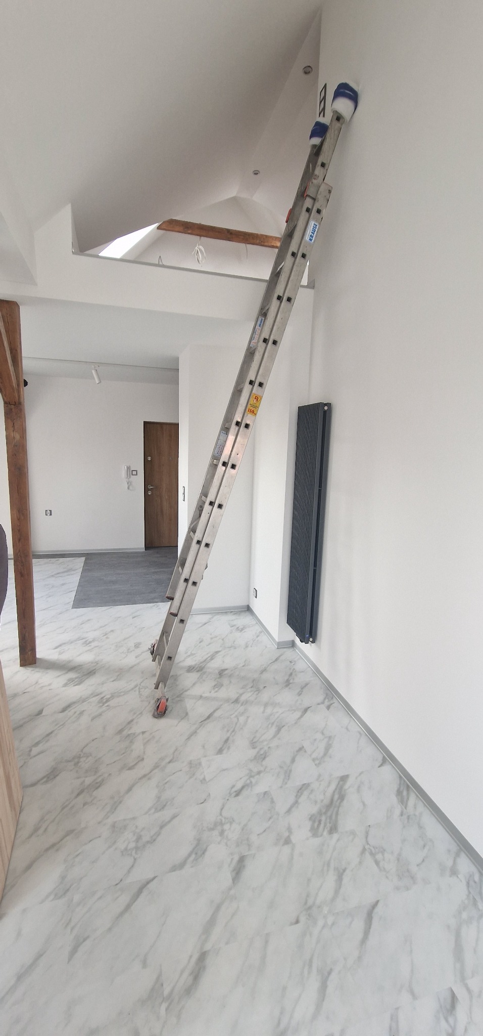 Aluminiowa drabina przystawiona do białej ściany w jasnym wnętrzu z marmurową podłogą, widoczny grzejnik panelowy i drewniany element konstrukcyjny pod sufitem.