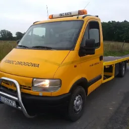 Żółta laweta z napisem 'POMOC DROGOWA', wyposażona w orurowanie ochronne z przodu, zaparkowana na asfaltowej drodze w pobliżu łąki.