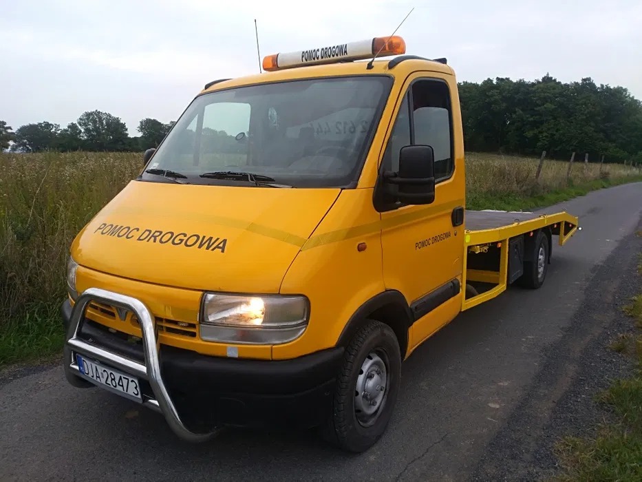Żółta laweta z napisem 'POMOC DROGOWA', wyposażona w orurowanie ochronne z przodu, zaparkowana na asfaltowej drodze w pobliżu łąki.