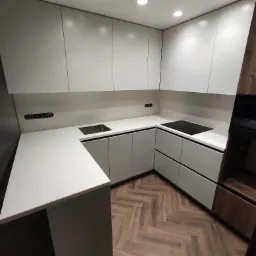 Nowoczesna kuchnia w kształcie litery U z białymi szafkami, białym blatem i drewnianą podłogą ułożoną w jodełkę; widoczny zlew i płyta indukcyjna.