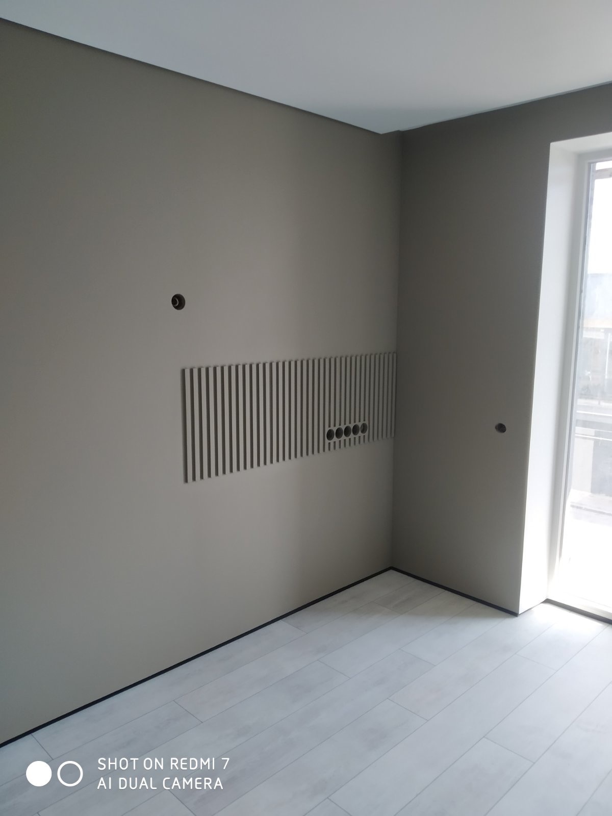 Pomalowane na szaro wnętrze pokoju z zamontowanymi listwami dekoracyjnymi na ścianie, ukrywającymi gniazdka elektryczne, oraz z jasną podłogą imitującą drewno.