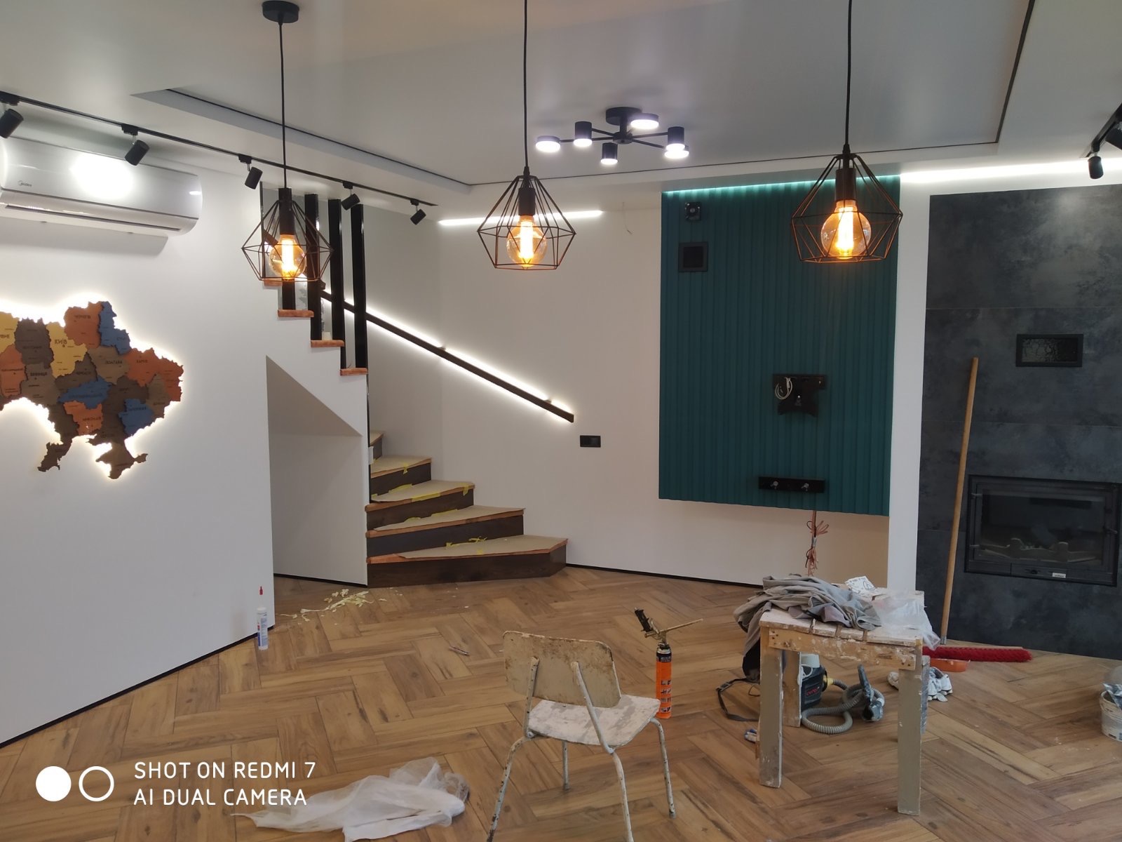 Nowoczesne wnętrze w trakcie wykańczania: parkiet w jodełkę, schody z podświetleniem LED, dekoracyjna mapa Ukrainy na ścianie, wiszące lampy w stylu industrialnym, ściana z panelami i fragment...