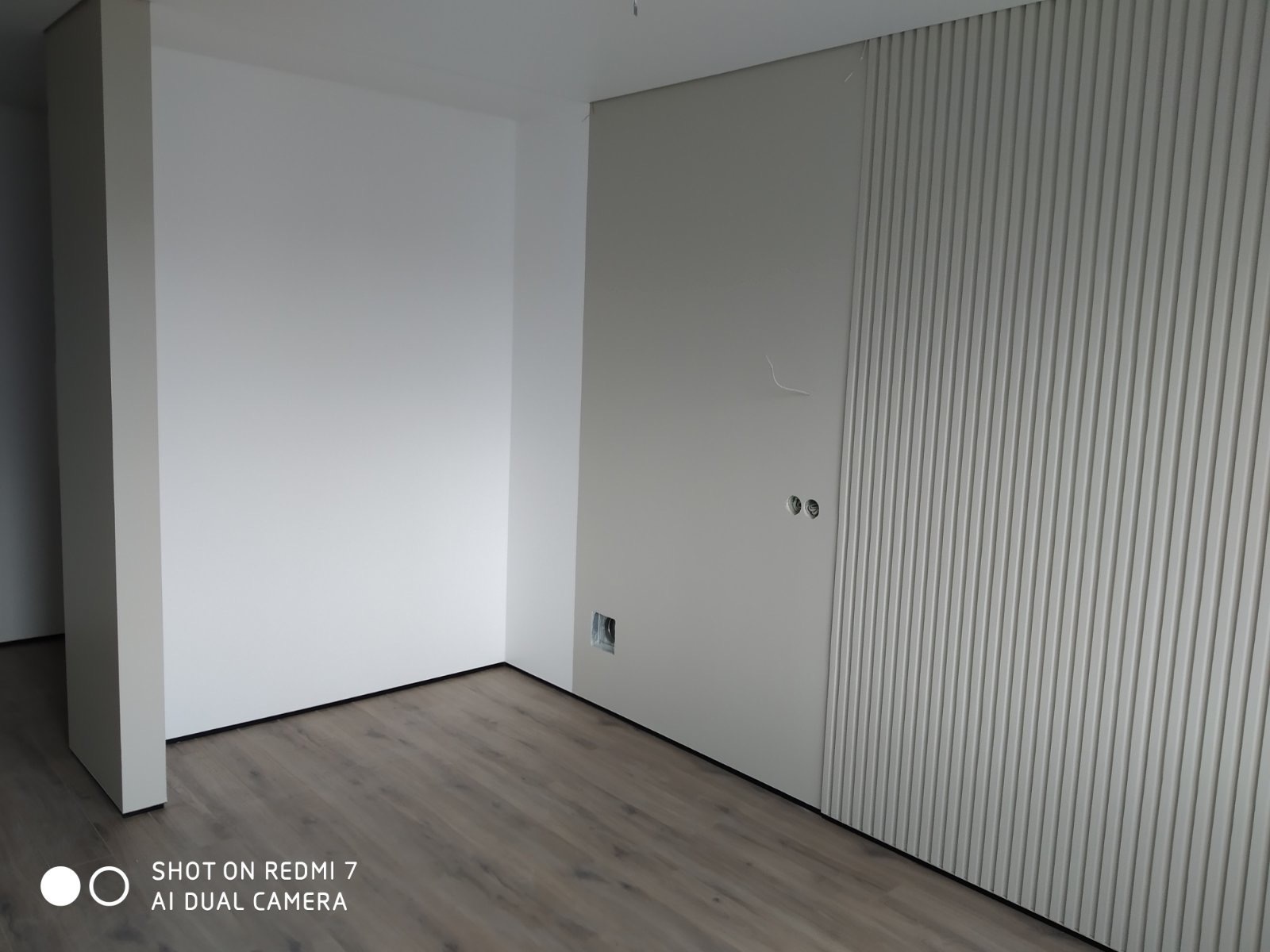 Wykończone wnętrze z panelami ściennymi w pionowe prążki, gładką ścianą, widocznymi gniazdkami elektrycznymi i drewnianą podłogą w odcieniach szarości.