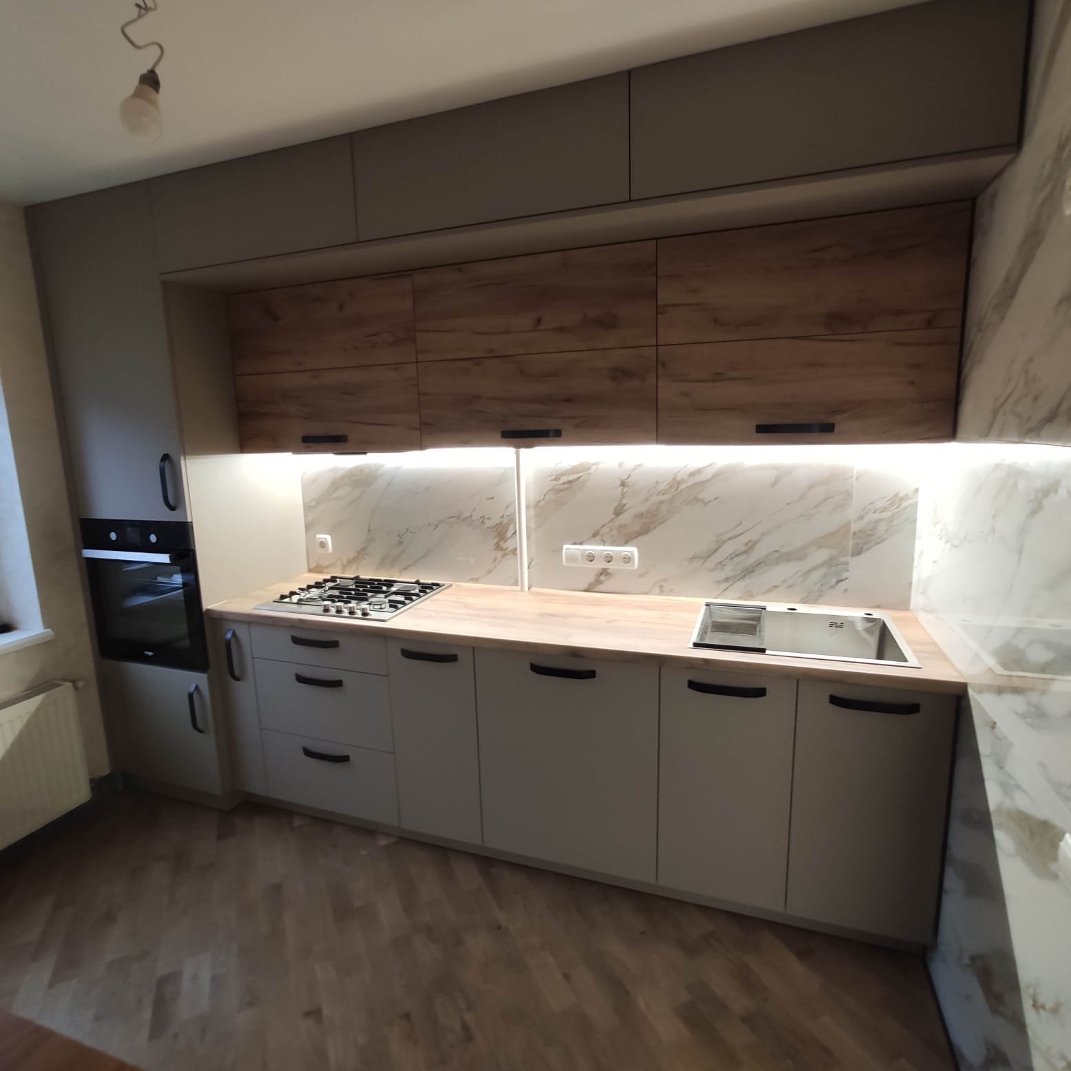 Nowoczesna kuchnia z szarymi szafkami dolnymi i górnymi w kolorze drewna, marmurowym backsplashiem i drewnianym blatem, oświetlona listwą LED pod szafkami wiszącymi.