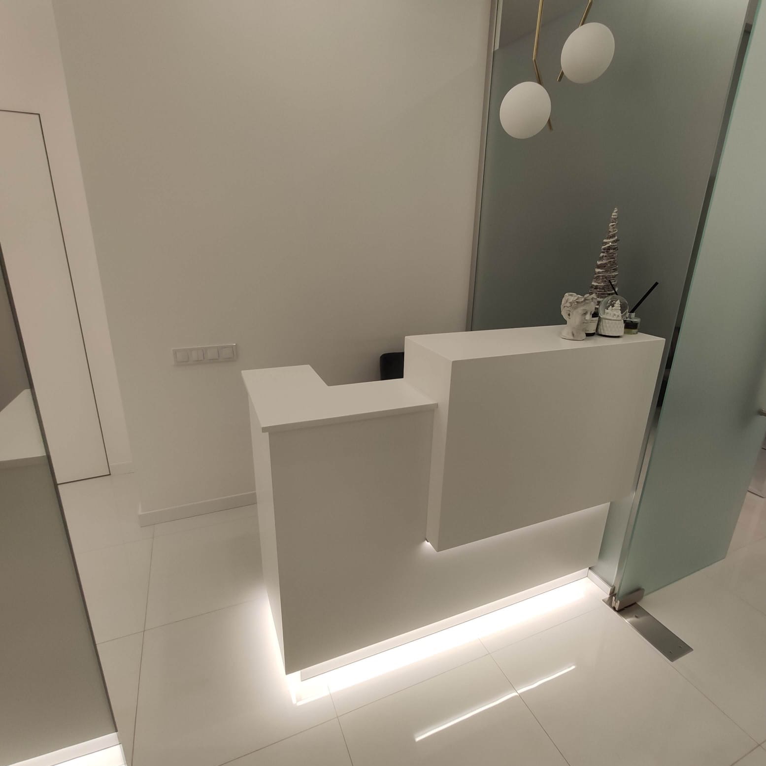 Minimalistyczna biała recepcja z podświetleniem LED u dołu, dekoracje na blacie: popiersie, szklana kula ze śniegiem i stożek, białe ściany i podłoga z płytek, szklane drzwi w tle.