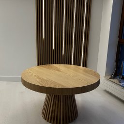 Usługi Stolarskie Ksawier Daniel Milewczyk - Okrągły, dębowy stół z żebrowaną podstawą, na tle ściany z drewnianych lameli z podświetleniem LED.