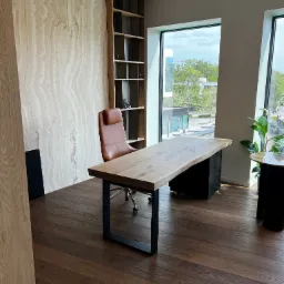 Biuro z dużym oknem, drewnianym biurkiem na metalowych nogach, skórzanym fotelem i drewnianą podłogą, widok na zieleń za oknem.