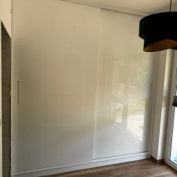 Usługi Stolarskie Ksawier Daniel Milewczyk - Biała szafa przesuwna z połyskiem, wbudowana we wnękę, z widocznymi drzwiami balkonowymi i fragmentem zielonego abażuru lampy.