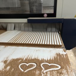 Usługi Stolarskie Ksawier Daniel Milewczyk - Frezarka CNC w trakcie obróbki płyty MDF z wyfrezowanymi rowkami, z rysunkami serc z wiórów na stole.