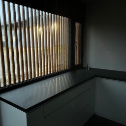 Usługi Stolarskie Ksawier Daniel Milewczyk - Fragment wnętrza z oknem przesłoniętym drewnianymi żaluzjami, blatem kuchennym z ciemnego kamienia i minimalistyczną zabudową szafek w odcieniach szarości.