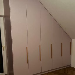 Usługi Stolarskie Ksawier Daniel Milewczyk - Szafa wnękowa z jasnoróżowymi frontami i drewnianymi uchwytami, dopasowana do skosu dachu na poddaszu.