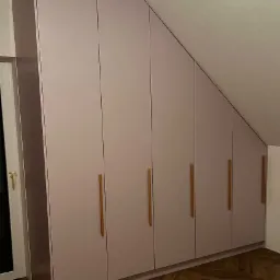Szafa wnękowa z jasnoróżowymi frontami i drewnianymi uchwytami, dopasowana do skosu dachu na poddaszu.