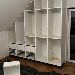 Usługi Stolarskie Ksawier Daniel Milewczyk - Biała szafa wnękowa z szufladami i półkami, zamontowana na poddaszu ze skosem, w trakcie montażu, widoczne prowadnice szuflad, osłonięta folią podłoga, brakujące drzwiczki szuflady obok.