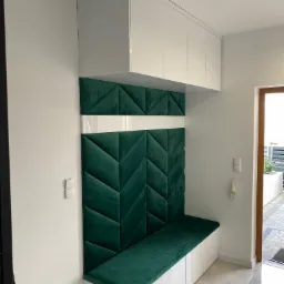 Wnęka z siedziskiem i szafkami w kolorze białym, z tapicerowanym panelem ściennym w kolorze butelkowej zieleni, ułożonym we wzór jodełki. Widoczny fragment podłogi z szarych płytek i drzwi wejściowe.