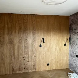 Drewniana zabudowa ściany z szafami, szufladami i ukrytymi drzwiami, oświetlona czarnymi kinkietami, na podłodze parkiet w jodełkę, obok ściana z tapetą w motywy ważek.