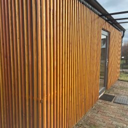 Usługi Stolarskie Ksawier Daniel Milewczyk - Nowoczesna elewacja budynku z pionowych, drewnianych listew w ciepłym odcieniu brązu, z widocznymi drzwiami wejściowymi i fragmentem chodnika z kostki brukowej.