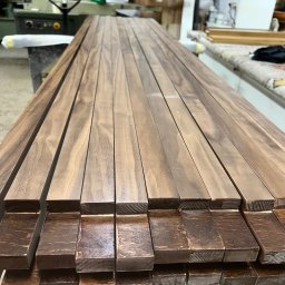 Usługi Stolarskie Ksawier Daniel Milewczyk - Przygotowane, lakierowane listwy drewniane ułożone w stos, widoczne słoje drewna i maszyny stolarskie w tle.