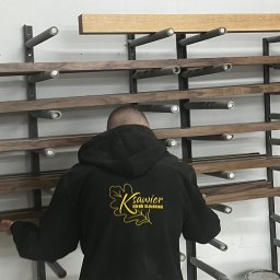 Usługi Stolarskie Ksawier Daniel Milewczyk - Pracownik w bluzie z logo firmy wybiera z regału drewniane listwy, przygotowane do dalszej obróbki.