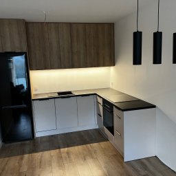 Usługi Stolarskie Ksawier Daniel Milewczyk - Nowoczesna, narożna zabudowa kuchenna w Luzinie z drewnopodobnymi szafkami górnymi, białymi szafkami dolnymi z czarnym blatem i wiszącymi, cylindrycznymi lampami.