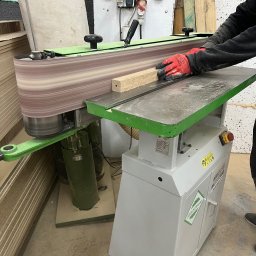 Usługi Stolarskie Ksawier Daniel Milewczyk - Obróbka drewnianego elementu na szlifierce taśmowej z zielonymi akcentami, operator w czerwonych rękawicach ochronnych, widoczna sterta płyt drewnianych w tle.