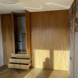 Usługi Stolarskie Ksawier Daniel Milewczyk - Drewniana szafa wnękowa z otwartymi drzwiami i wysuniętymi szufladami, widoczne półki wewnątrz, na ścianie tapeta z motywem roślinnym, podłoga parkiet w jodełkę, wystające kable elektryczne.