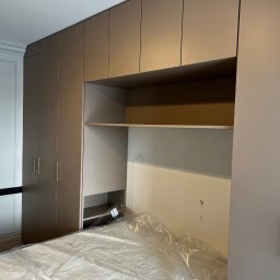 Usługi Stolarskie Ksawier Daniel Milewczyk - Beżowa zabudowa meblowa z szafami i półkami nad łóżkiem przykrytym folią ochronną, widoczne złote uchwyty szafek i fragment białej sztukaterii.