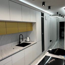 Usługi Stolarskie Ksawier Daniel Milewczyk - Nowoczesna kuchnia z białymi szafkami, złotymi akcentami i czarną armaturą, widok w głąb korytarza z toaletą.