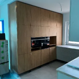 Wysoka zabudowa kuchenna z drewnianymi frontami w odcieniach beżu, z wbudowanymi piekarnikami, w trakcie montażu, widoczne narzędzia i części wyposażenia.