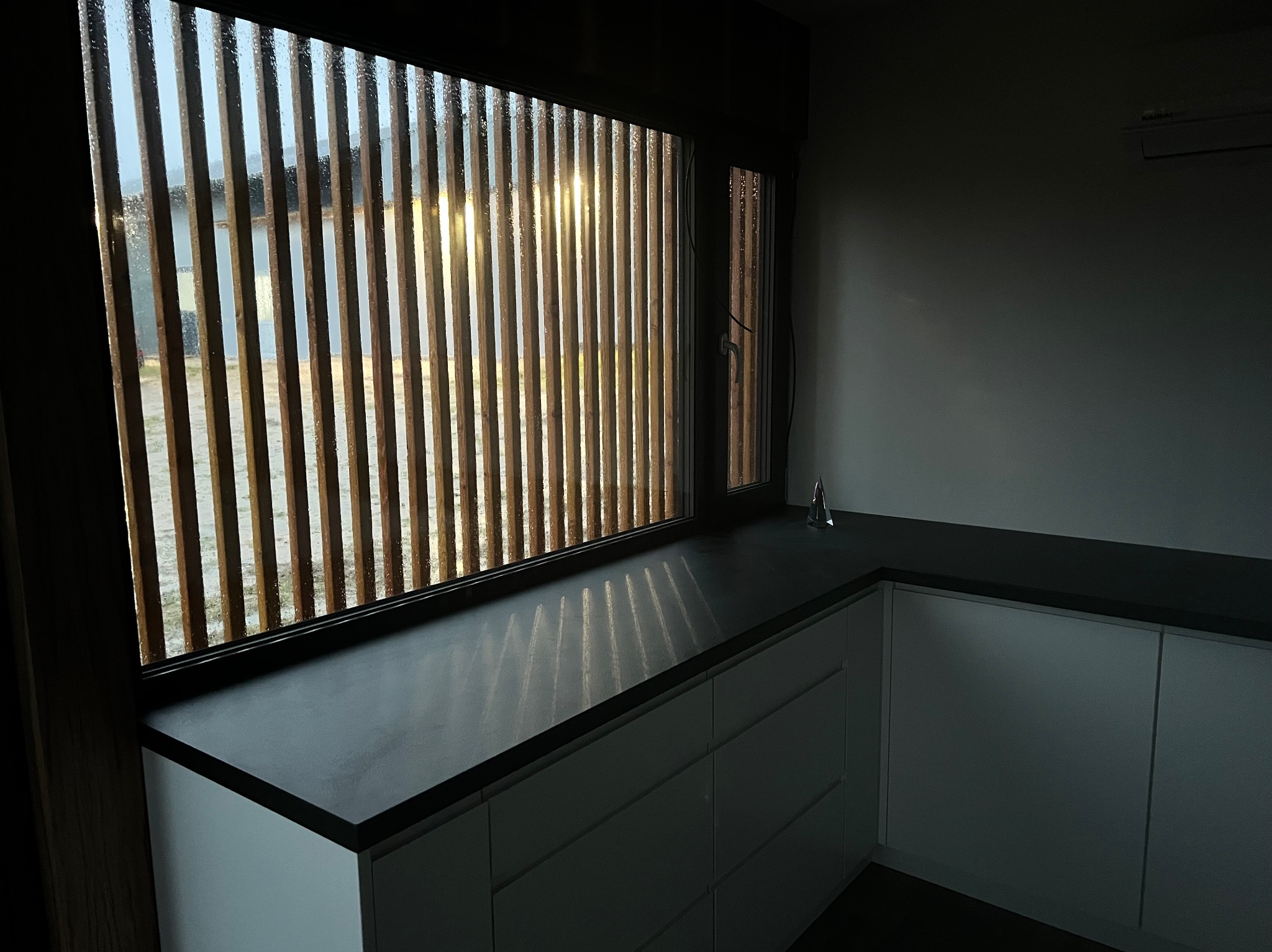 Fragment wnętrza z oknem przesłoniętym drewnianymi żaluzjami, blatem kuchennym z ciemnego kamienia i minimalistyczną zabudową szafek w odcieniach szarości.