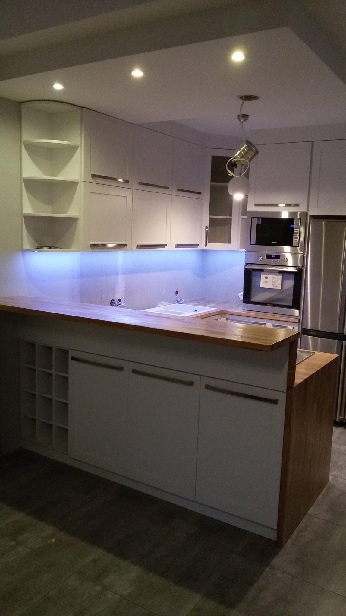 Nowoczesna kuchnia z białymi szafkami z drewnianym blatem, podświetleniem LED i otwartymi półkami na wino, w tle sprzęt AGD ze stali nierdzewnej.